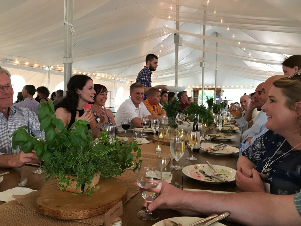 Group of people enjoying a meal at a corporate event table in a tent, featuring greenery centerpieces and a relaxed atmosphere, emphasizing connection and engagement at On Sunny Slope Farm.